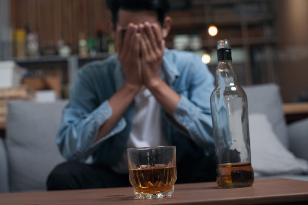 Man with head in hands behind a glass and bottle of whisky on a table in a living room