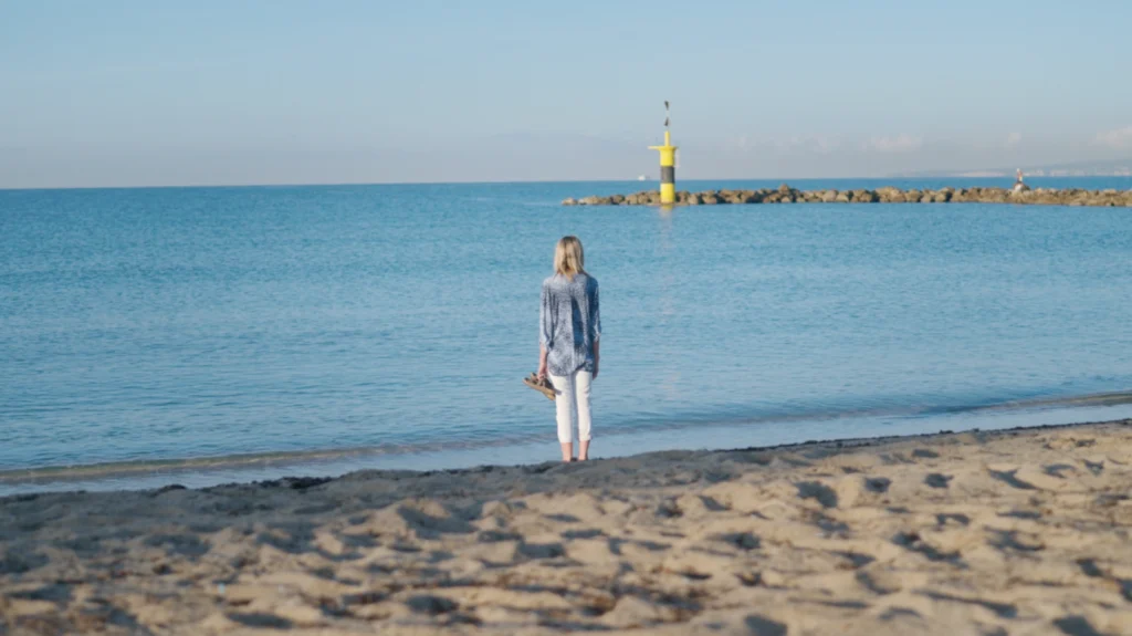 Person standing on a sandy beach looking out at the calm sea toward a yellow beacon on a breakwater