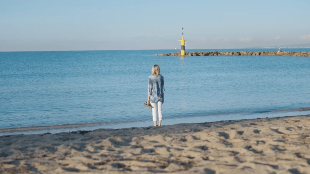 Person standing on a sandy beach looking out at the calm sea toward a yellow beacon on a breakwater