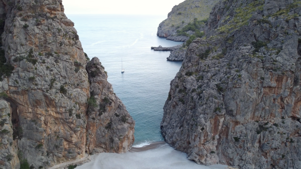 Sailboat in a narrow sea cove between steep rocky cliffs, leading to a small pebble beach below.